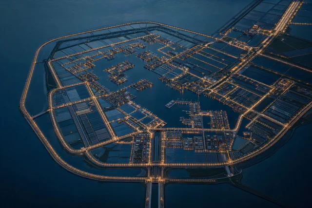 Aerial view at dusk of a large artificial island ringed by a lit road, with interior basins, marinas, blocks of buildings and a bridge leading off at the bottom.