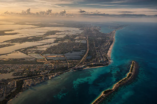 Aerial view at sunset of a low-lying coastal city along a sandy shoreline, with wetlands and shallow ponds inland, turquoise water offshore and a narrow sandy spit in the foreground.