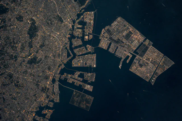 Aerial view of a dense city at left and rectangular reclaimed port and industrial islands extending into a dark bay at right.