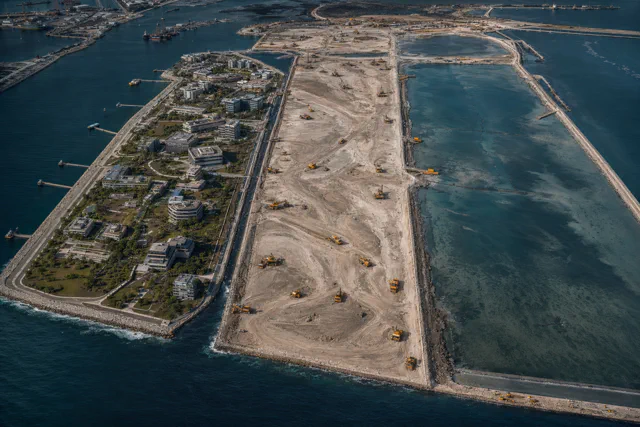 Aerial view of a reclaimed peninsula, with landscaped buildings on one side, a graded sand platform covered in construction equipment in the middle and a shallow basin beside it.